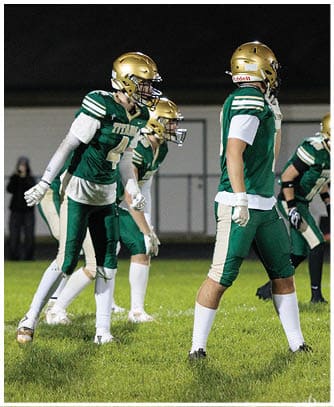 Three people in green and gold football uniforms stand on a grassy field, appearing ready for action during a nighttime game. Description generated by AI