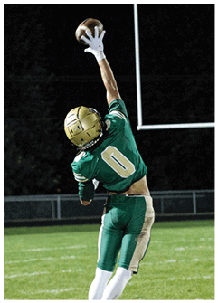 A person wearing a green sports jersey and white gloves is reaching up to catch a football against a dark background. Description generated by AI