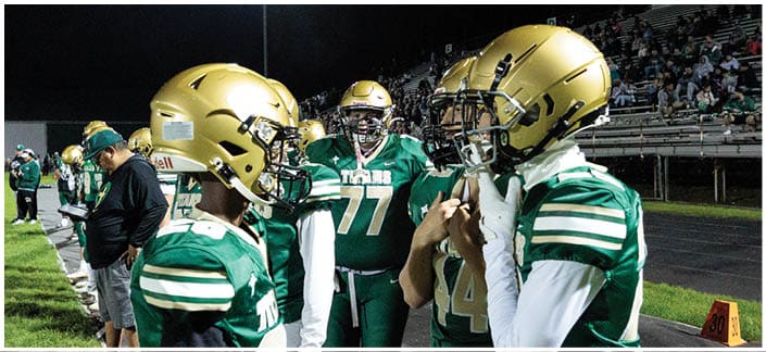 Three people wearing green and white football uniforms and gold helmets are standing and talking on the sidelines of a nighttime football game. Description generated by AI
