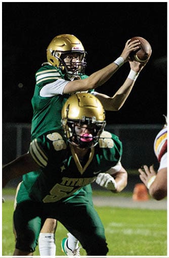 A person in a green uniform is preparing to throw a football while another person in a white and yellow uniform is positioned defensively in front of them on a nighttime football field. Description generated by AI