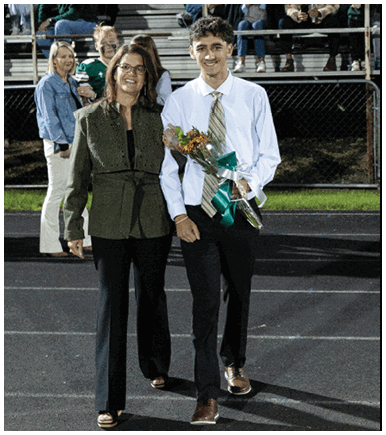 A person wearing glasses and a green jacket is smiling and standing on a track field while holding hands with another person partially visible in a white shirt, with other people and bleachers in the background. Description generated by AI