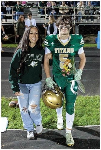 A person in a green football uniform holding a helmet and flowers walks arm-in-arm with another person wearing a green sweatshirt and ripped jeans on a sports field with spectators in the background. Description generated by AI