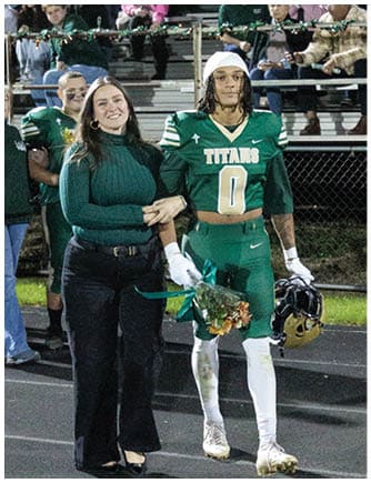 An adult in a green football uniform holding a helmet and flowers walks arm-in-arm with another adult wearing a green sweater and black pants on a track with spectators in the background. Description generated by AI