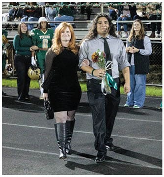 A person in a dress shirt and tie holding flowers stands arm-in-arm with another person in a black top on a sports field with spectators seated in bleachers behind them. Description generated by AI