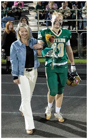 A person in a green football uniform with face paint is holding a bouquet of flowers and standing on a field with spectators seated in the background. Description generated by AI