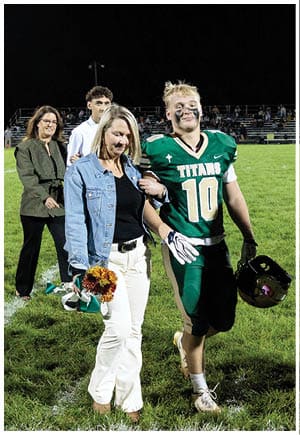 People standing on a grassy sports field with white boundary lines, some holding flowers and one holding a football helmet. Description generated by AI
