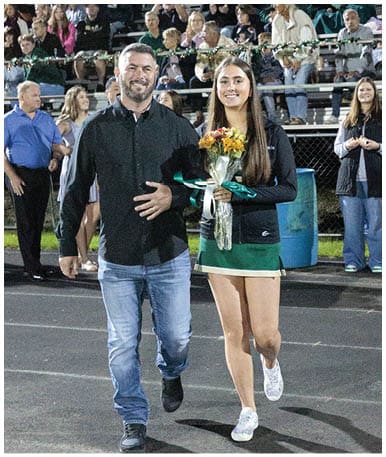 A person with long straight hair wearing a dark jacket is smiling and holding a bouquet of flowers in front of a crowd seated on bleachers decorated with garlands. Description generated by AI
