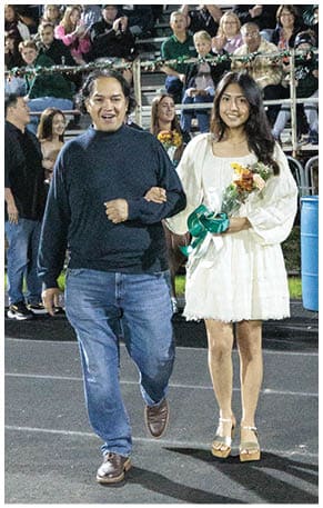 A person with long dark hair wearing a white dress holds a bouquet of flowers with a green ribbon while standing in front of seated people in bleachers at an outdoor event. Description generated by AI