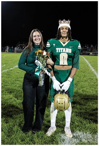A person wearing a football uniform and crown stands on a field holding a helmet while another person beside them holds a bouquet of flowers and smiles. Description generated by AI