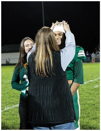A person is placing a crown on another person wearing a green sports uniform on a grassy field at night while two other people stand nearby holding flowers and smiling. Description generated by AI