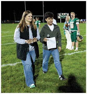 People standing on a football field at night with some holding flowers and helmets while one person reads from a paper. Description generated by AI
