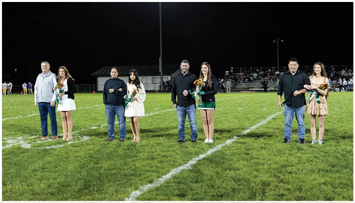 People standing on a grassy field with white painted lines and numbers, some holding football helmets. Description generated by AI