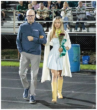 A person wearing glasses and a blue pullover stands with arms crossed on a sports field with bleachers and spectators in the background. Description generated by AI