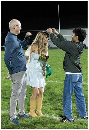 Two people stand on a grassy field with one holding a bouquet of flowers and wearing a light blue dress and yellow boots while the other wears gray pants, blue shoes, and a blue long-sleeve shirt. Description generated by AI