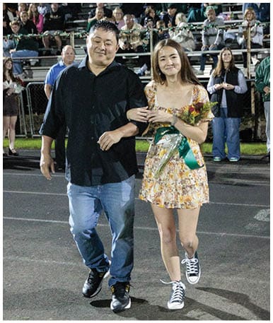 A person wearing a floral dress and holding a bouquet of flowers stands in front of a crowd seated on bleachers decorated with string lights. Description generated by AI