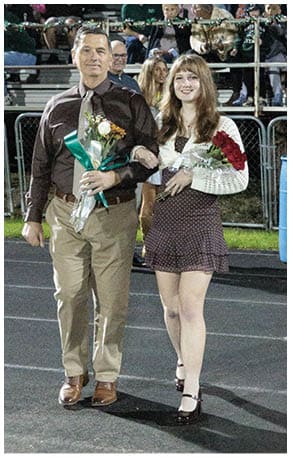 A person with long hair wearing a white sweater and holding a bouquet of red roses is smiling in front of a crowd seated on bleachers at an outdoor event. Description generated by AI