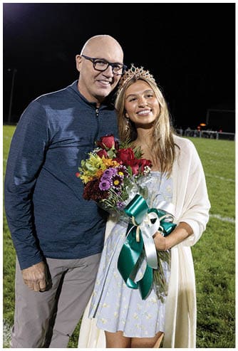 An adult wearing glasses and a long-sleeve shirt stands next to a smiling person wearing a crown, a light blue dress, and a white shawl while holding a bouquet of flowers on a grassy field at night. Description generated by AI