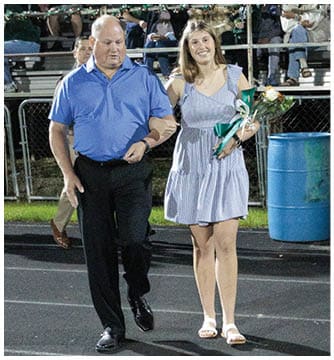 An adult in a blue shirt is walking arm-in-arm with a smiling adult in a striped dress holding a bouquet of flowers in front of a seated crowd at an outdoor event. Description generated by AI