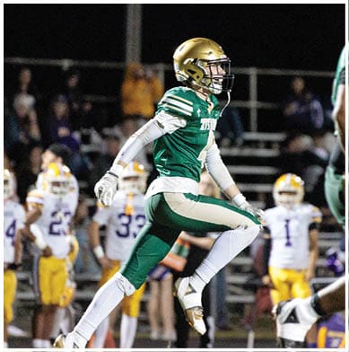A person in a green and gold football uniform is running on a field with other players and spectators in the background during a nighttime game. Description generated by AI