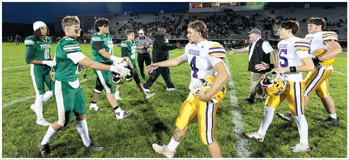 People in football uniforms from two teams are shaking hands on a grassy field under stadium lights at night with spectators in the background. Description generated by AI