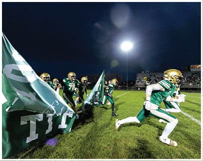 A group of football players in green and gold uniforms run onto the field through large green flags held by people at night under bright stadium lights. Description generated by AI