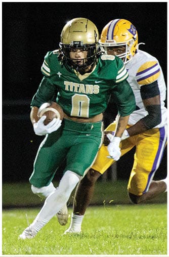 A person in a green football uniform with a gold helmet is running with a football while another person in white reaches out to try to tackle them on a grassy field at night. Description generated by AI