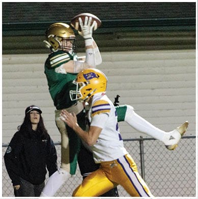 A person in green and gold football gear is jumping to catch a football while another person in white and yellow football gear runs nearby on a field with a chain-link fence and building in the background. Description generated by AI