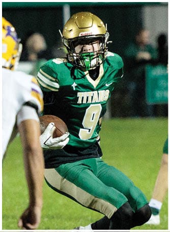 A person wearing a green football uniform with the word Titans and number 9 is running on a grassy field while holding a football. Description generated by AI