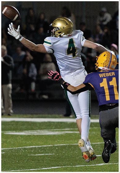 Two people in football uniforms are engaged in a play on a field, with one person in white and green jumping while the other in purple and yellow reaches out to them. Description generated by AI