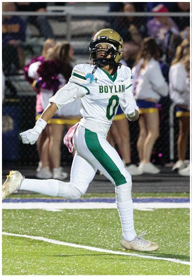 A person wearing a white and green football uniform with the name “Boylan" and number zero is running on a football field with cheerleaders in the background. Description generated by AI