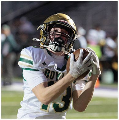 A person wearing a football helmet and jersey is catching a football during a game. Description generated by AI