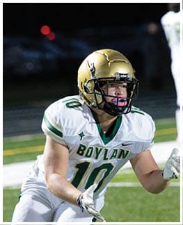 A person wearing a football helmet and white jersey with green and gold accents is on a field, with another person in white athletic clothing blurred in the background. Description generated by AI