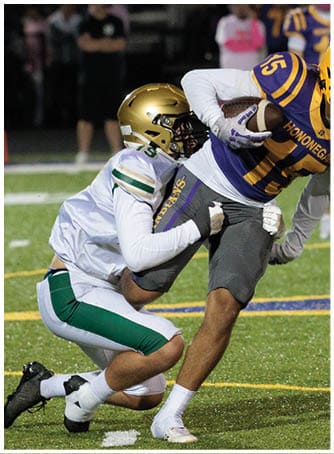 A person in a purple and yellow football uniform is running with the ball while being tackled by another person in a white and green uniform on a football field. Description generated by AI