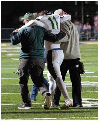Four people walk together on a football field, with one person in a sports uniform being supported by two others. Description generated by AI