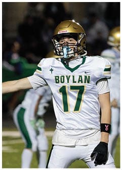A person wearing a gold football helmet and a white jersey with green and yellow accents is extending one arm on a football field during a game. Description generated by AI