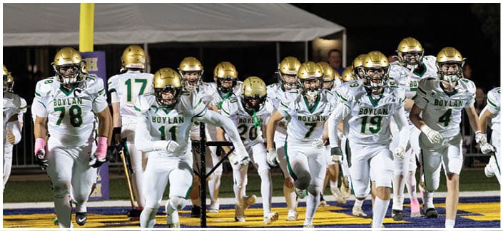 A group of football players in white and green uniforms with gold helmets are running together on a field under stadium lights. Description generated by AI