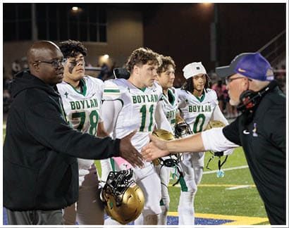 Four people in football uniforms with “Boylan" on the jerseys stand on a field while two others in casual clothing extend their hands toward each other. Description generated by AI