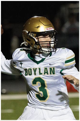 A person wearing a gold football helmet and a white jersey with “BOYLAN" and the number 3 is preparing to throw a football on a field. Description generated by AI