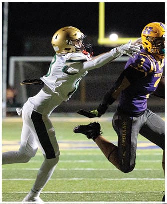 Two people in football uniforms are actively reaching for the ball on a lit field during a nighttime game. Description generated by AI