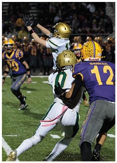 Two people in white and gold football uniforms are engaged in a play while two people in purple and yellow uniforms are nearby on a football field during a game. Description generated by AI