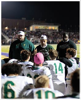 A group of adults in sportswear are coaching and talking to a team of players wearing white and green football uniforms on a field at night. Description generated by AI