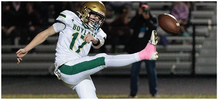 A person wearing a football uniform with a gold helmet and a pink sock is kicking a football on a field with blurred spectators in the background. Description generated by AI