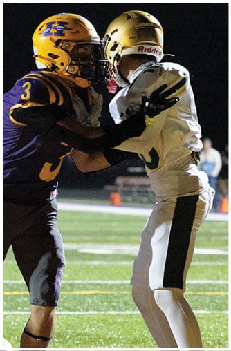 Three football players in action on a field at night with one player in white tackling a player in purple and yellow while another player in purple and yellow runs nearby holding the ball. Description generated by AI