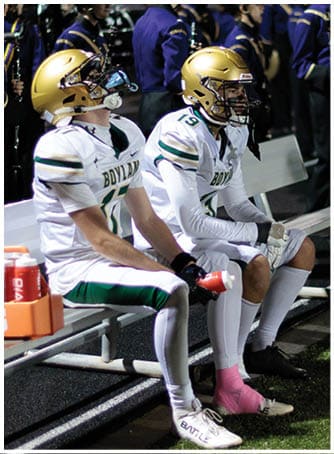 People in football uniforms sit on a bench near a marching band dressed in dark uniforms with yellow plumes on their hats at a nighttime outdoor event. Description generated by AI