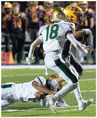 Two people in white and green football uniforms are tackling a person in a purple and yellow football uniform on a field with other players in the background. Description generated by AI