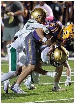 Two people in football uniforms are tackling a third person in a football uniform who is holding a football on a grassy field during a game. Description generated by AI