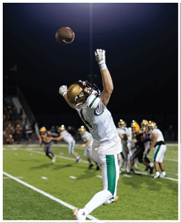 A person wearing a gold helmet and white sports uniform with green and gold stripes is reaching up with both hands toward a football in midair against a dark background. Description generated by AI