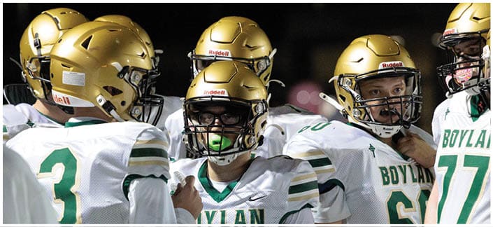 A group of football players wearing white and green uniforms with gold helmets are gathered closely together, appearing to be in a huddle during a game. Description generated by AI