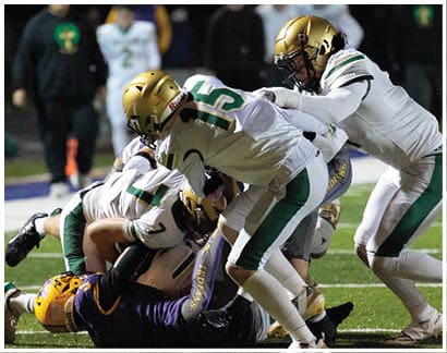 Several football players in white and green uniforms tackle a player in a purple and yellow uniform on a field during a game. Description generated by AI