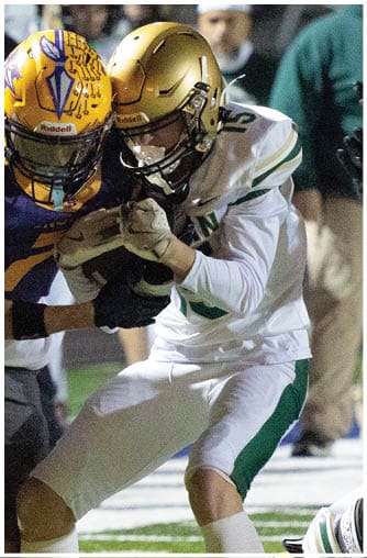 Two people in football uniforms are engaged in a close tackle on a field with others and a coach visible in the background. Description generated by AI